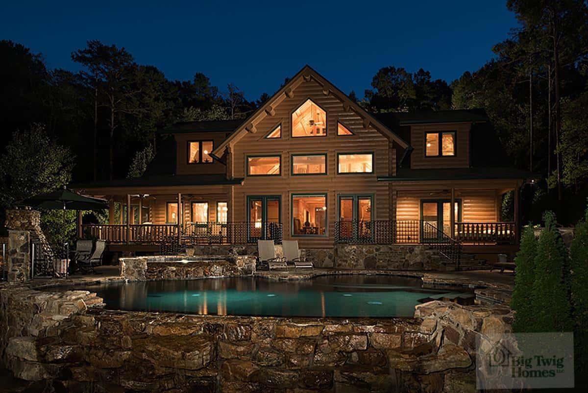 Luxury Bennett cedar log home at dusk with illuminated windows, stonework, and pool